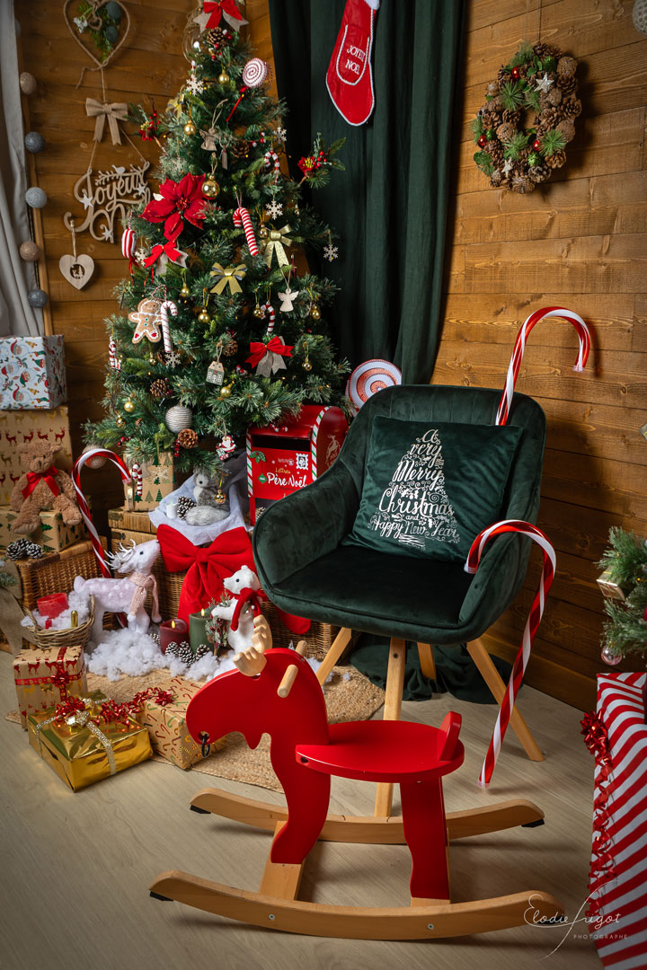 Photo du décor traditionnel des mini-séances de Noël avec fauteuil vert renne à bascule sapin cadeaux sur fond bois au Studio Elodie Frigot Photographe